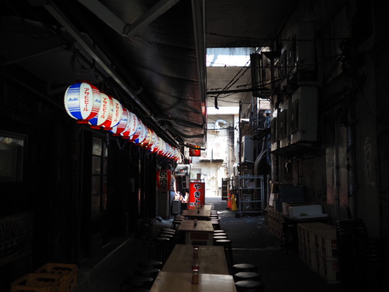 アメ横の薄暗い路地裏、簡易的な椅子とテーブルが並んだ、居酒屋の提灯が並び赤い焼き鳥の看板がある風景